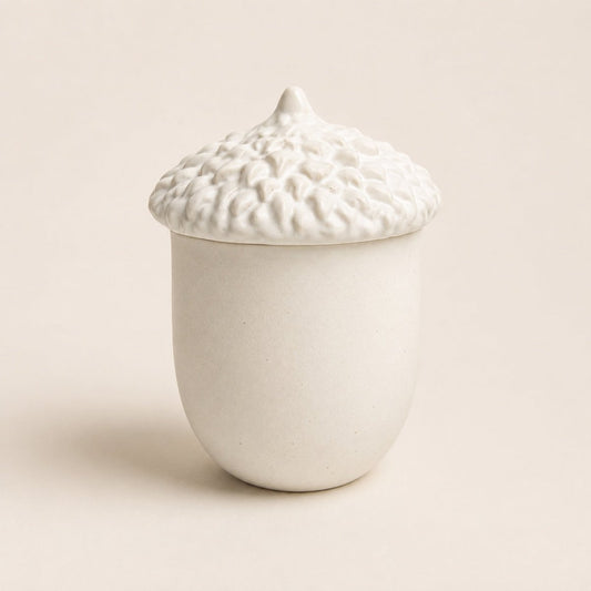 A small white stone acorn-shaped container with a textured, ridged lid and a smooth rounded body, set against a solid warm beige background.