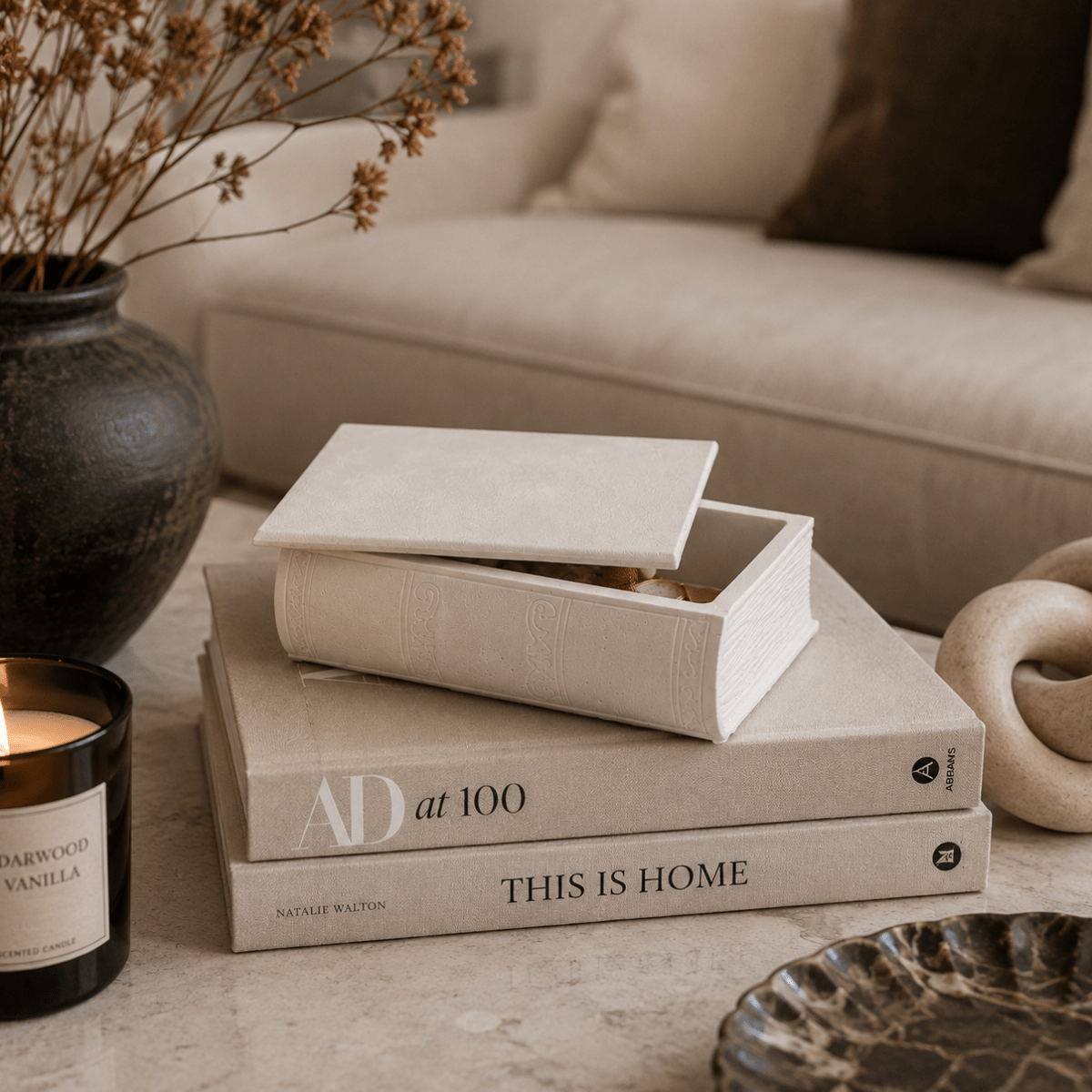 A cream-colored textured book-shaped jewelry box with its lid slightly open, styled on top of two hardcover books on a light stone surface in a cozy living room setting.