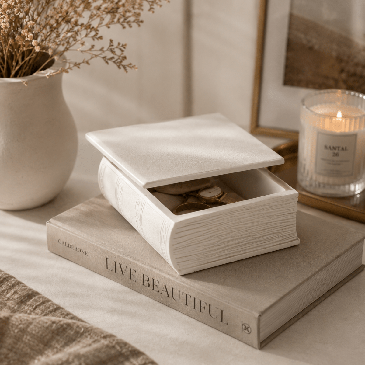 A white Jesmonite storage box designed to look like an open book, with its lid propped open, resting on a decorative book. The box contains gold-toned jewellery, including a watch and earrings.