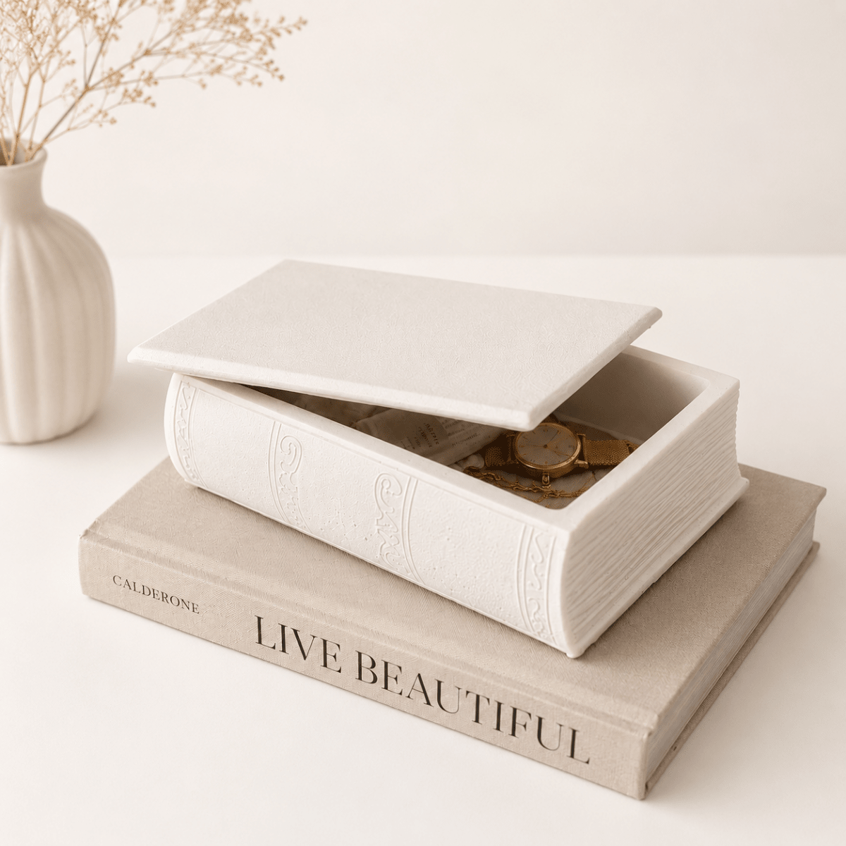 A white book-shaped jewellery box with a lid, styled on top of a beige book with the words 'LIVE BEAUTIFUL' on its spine. A small white vase with dried stems sits in the background on a clean, light-colored surface.