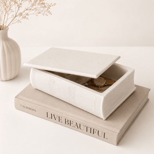 A white book-shaped jewellery box with a lid, styled on top of a beige book with the words 'LIVE BEAUTIFUL' on its spine. A small white vase with dried stems sits in the background on a clean, light-colored surface.