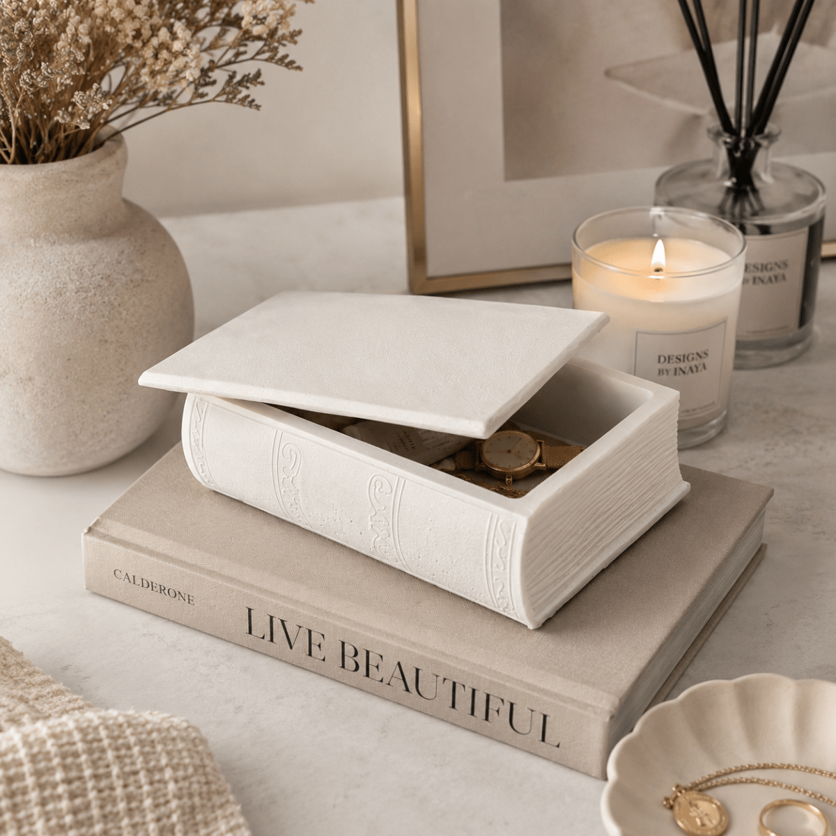 A white book-shaped stone Trinket box with its lid propped open sits on a beige book on a light marble surface. The box contains gold-toned jewellery, including a watch and rings.