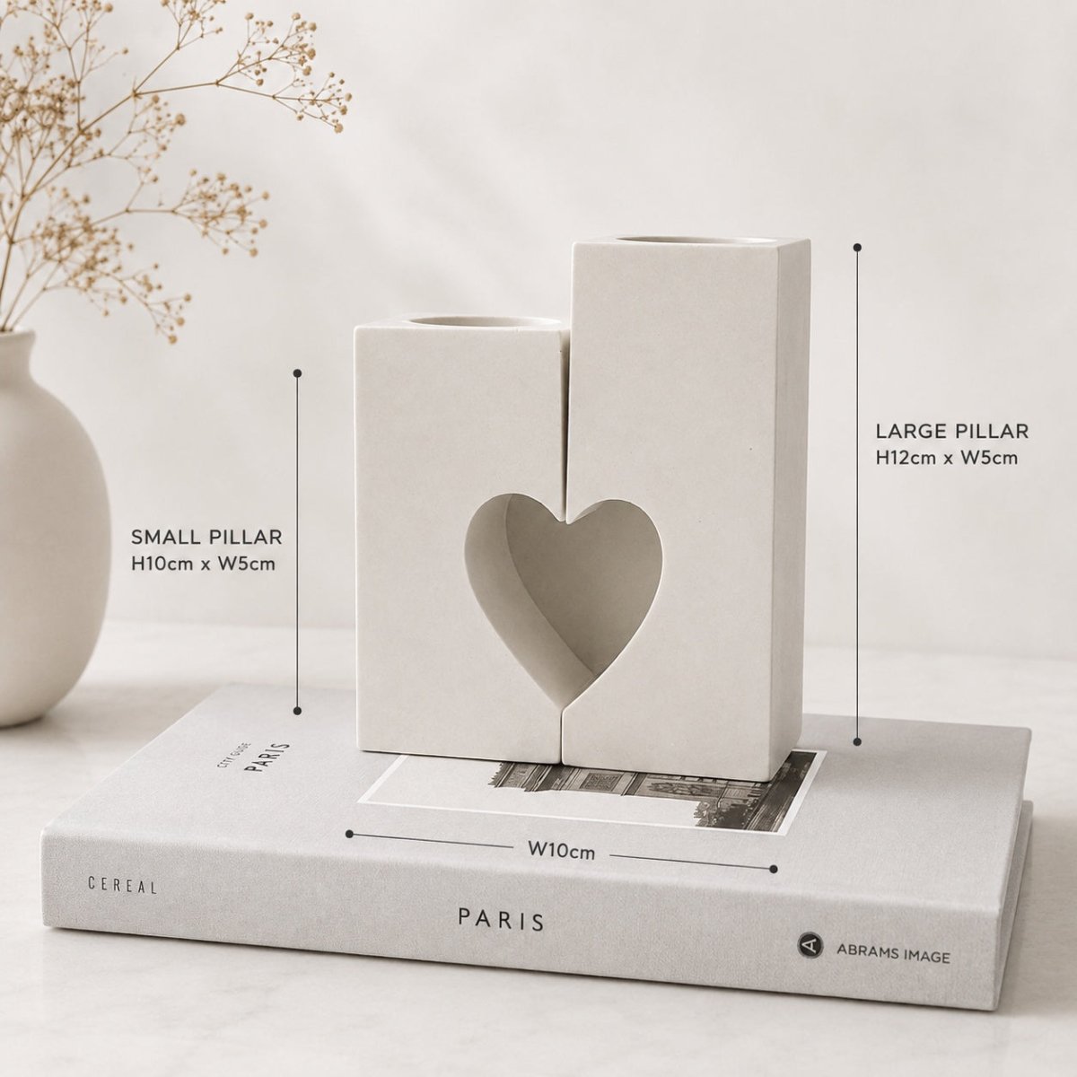 Two minimalist white Handcrafted stone candle holders shaped like pillars, with one featuring a heart-shaped cutout, displayed on top of a book with dimension callouts showing measurements.