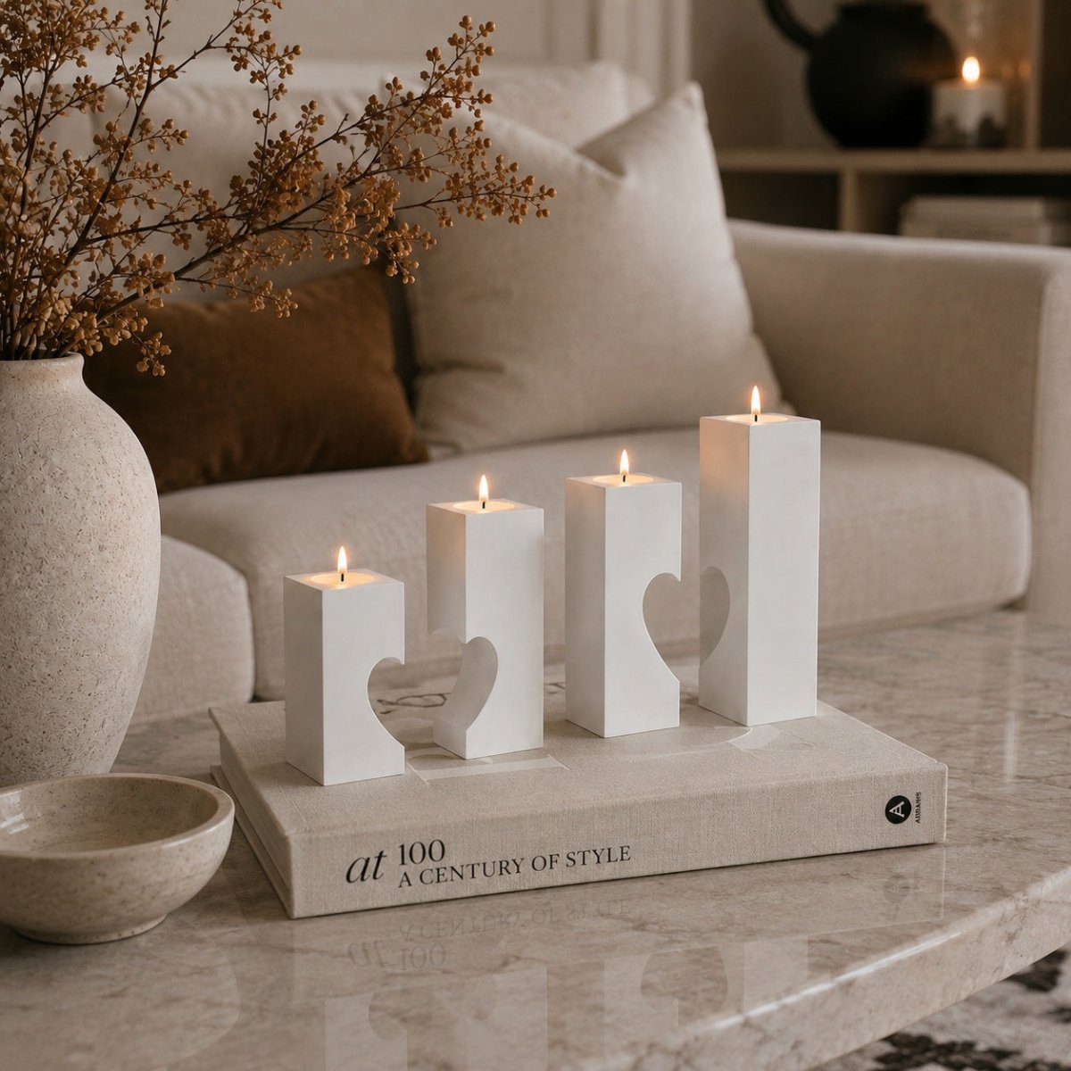 Four white geometric candle holders with heart-shaped cutouts, lit and arranged on a book atop a marble coffee table in a cozy living room setting. styled for modern home decor