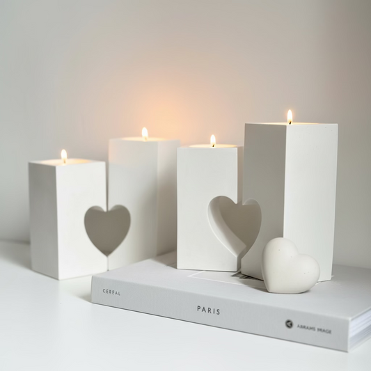 Four white rectangular pillar-style candle holders with heart-shaped cutouts, arranged on a white surface with candles lit and a small white heart-shaped object beside them. A book labeled PARIS sits underneath.