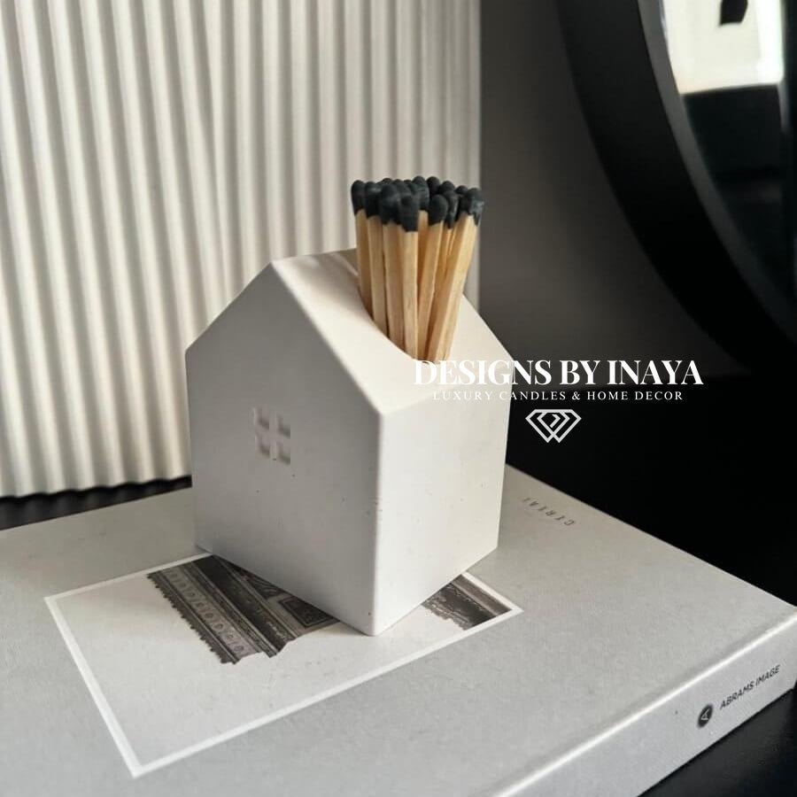 Matches in a white house-shaped container on a surface with 'Designs by Inaya' branding.