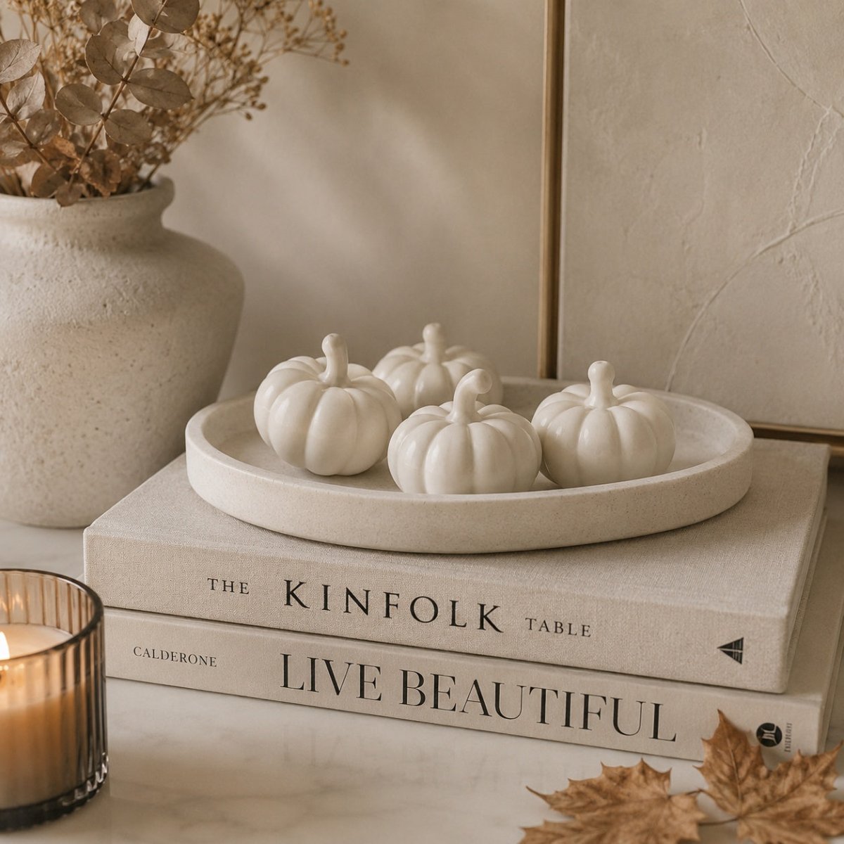 Four small white handcrafted stone pumpkin ornaments arranged on a round beige tray, which sits on top of two stacked books. The scene is styled with a textured ceramic vase containing dried branches, a lit candle in a glass holder, and dried autumn leaves on a light surface.